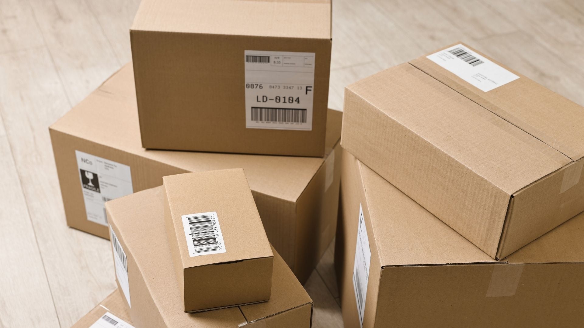 Ein Stapel aus mehreren braunen Versandkartons in unterschiedlichen Größen liegt auf einem hellen Holzboden. Auf den Kartons sind weiße Versandetiketten mit deutlich sichtbaren Barcodes und Aufdrucken angebracht.